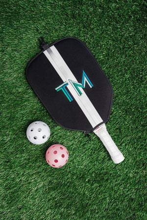 A black and silver paddle cover is customized with an embroidered monogram and placed next to some pickleballs