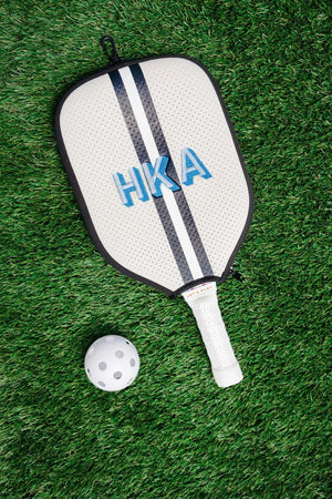 A white and navy paddle cover is customized with an embroidered monogram and placed next to a pickleball