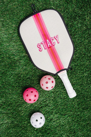 A pink and white paddle cover is customized with an embroidered name and placed next to some pickleballs