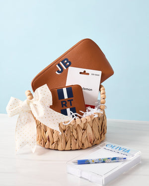 A basket is filled with a tan leather pouch and a tan jewelry case with navy and white monograms
