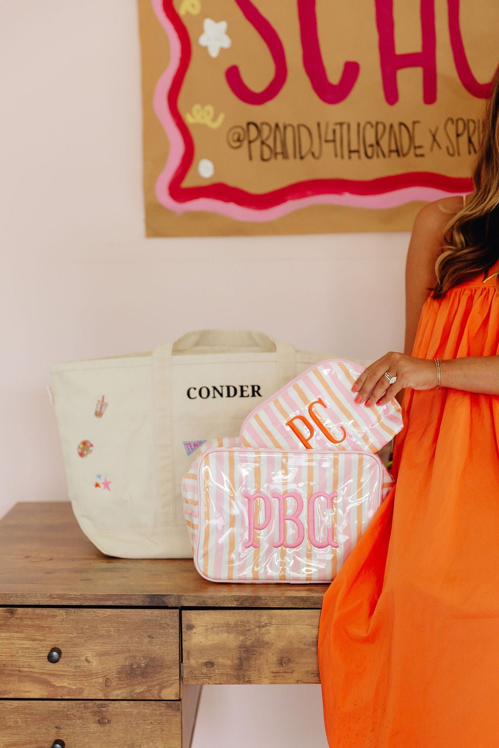 Cream canvas tote bag embroidered 'CONDER' in pink, with school icon patches (iced coffee, palette, star, teach pennant, books, school bus, crayons), paired with a pink and orange striped cosmetic pouch monogrammed 'PBC', on a white background.