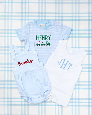Rainey Laine baby outfit trio featuring a Short Sleeve Striped Bubble in light blue with green tractor trailer motif and personalized name ‘Henry,’ a light blue suspender bubble with red script name ‘Brooks,’ and a white seersucker bubble with blue monogram ‘JHT,’ all laid out on a soft blue plaid background.