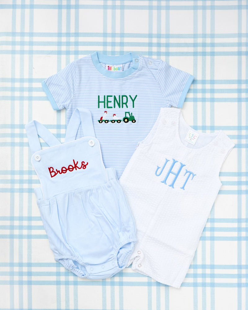 Rainey Laine baby outfit trio featuring a Short Sleeve Striped Bubble in light blue with green tractor trailer motif and personalized name ‘Henry,’ a light blue suspender bubble with red script name ‘Brooks,’ and a white seersucker bubble with blue monogram ‘JHT,’ all laid out on a soft blue plaid background.