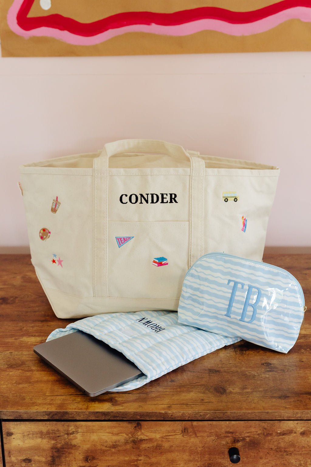 Canvas teacher tote bag embroidered 'CONDER' with school-themed icon patches, paired with a pink and orange striped laptop sleeve and matching cosmetic pouch monogrammed 'PBC'.