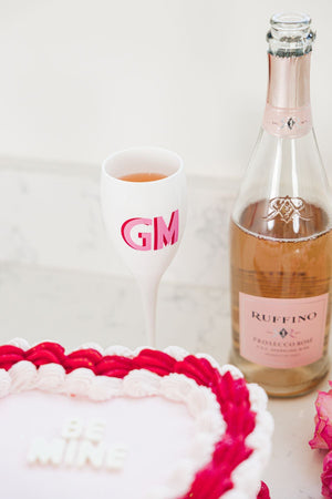 A white champagne flute with a pink and red monogram sits next to a bottle of wine and a "be mine" cake