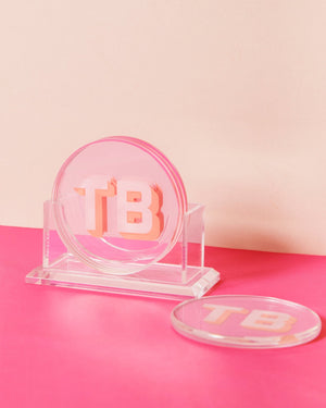 A coaster set sits in their stand with a pink and melon monogram against a bright pink background.