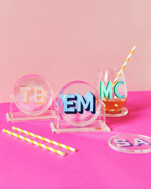 Two sets of coasters customized with pink and blue monograms sit in their stand next to a glass of wine.