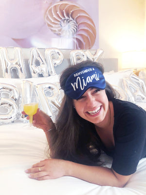 A woman lays on a bed decorated with birthday balloons while wearing our Miami-themed sleep mask