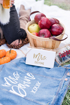 An acrylic clutch that reads "Mrs. Devine" sits next to a custom denim jacket and barrel of apples