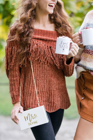 A woman in a rust-colored sweater sports a clutch that reads "Mrs. Devine"