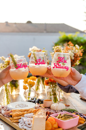 Three people cheers with their wine glasses that are personalized with their names in a pink font.