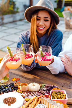Three people cheers with their wine glasses that are personalized with their names in a pink font.
