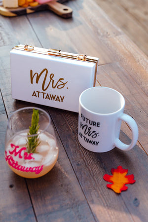 A wine glass, white mug, and a white clutch are all personalized for a bride.
