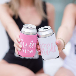A white can cooler that reads "the bride" in pink and a pink koozie that reads "bride's tribe" come together to cheers.