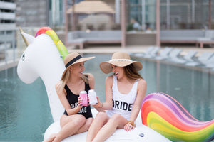 A bride and her friend cheers their drinks with can coolers in the pool wearing floppy beach hats and customized swimsuits.