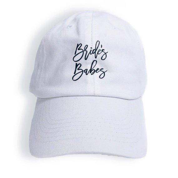 A pink baseball hat which says "Bride's Babes" in script white font sits next to a white baseball hat which says "Bride" in a pink script font.