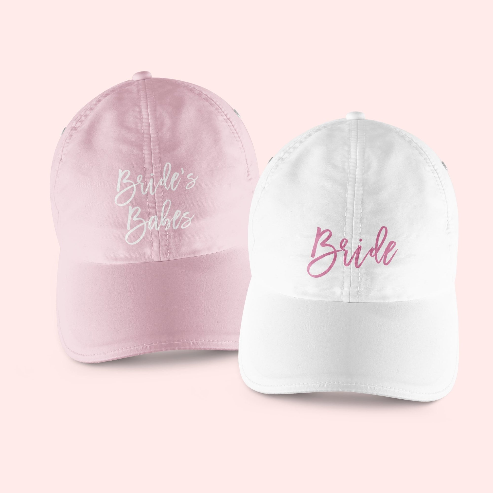 A pink baseball hat which says "Bride's Babes" in script white font sits next to a white baseball hat which says "Bride" in a pink script font.