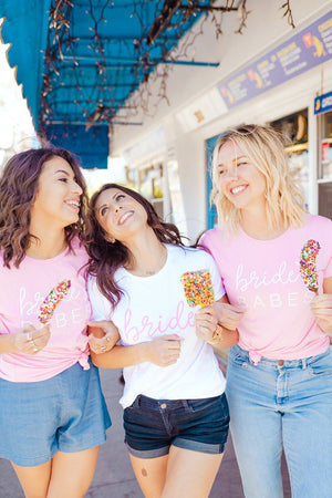 A bride and her two friends hold arms and laugh as they are styled in "Bride" and "Bride's Babes" Shirts.