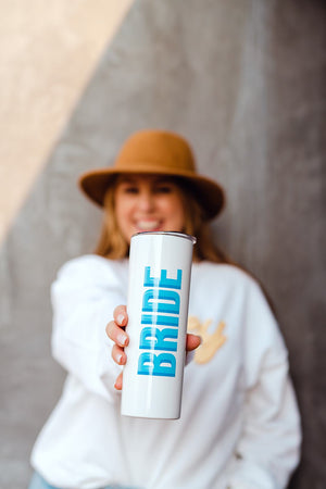 A woman holds out a white tumbler that is customized with blue text that says "Bride"