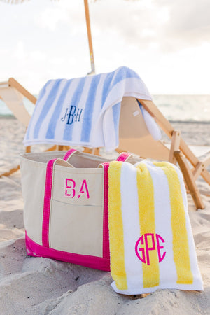 A pink canvas tote is placed on the beach with a blue and yellow cabana towel that are embroidered with monograms.
