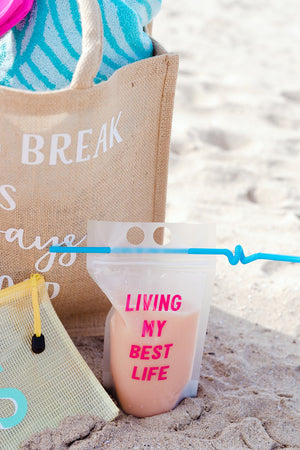 A party pouch sits in the sand with some other beach supplies and reads "Living my best life."