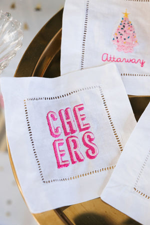 A cocktail napkin with "Cheers" embroidered on it in pink thread colors.
