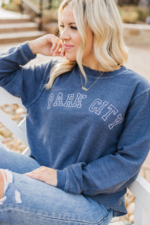A girl sits on a bench showing off her navy corded sweatshirt which reads "Park City" in white