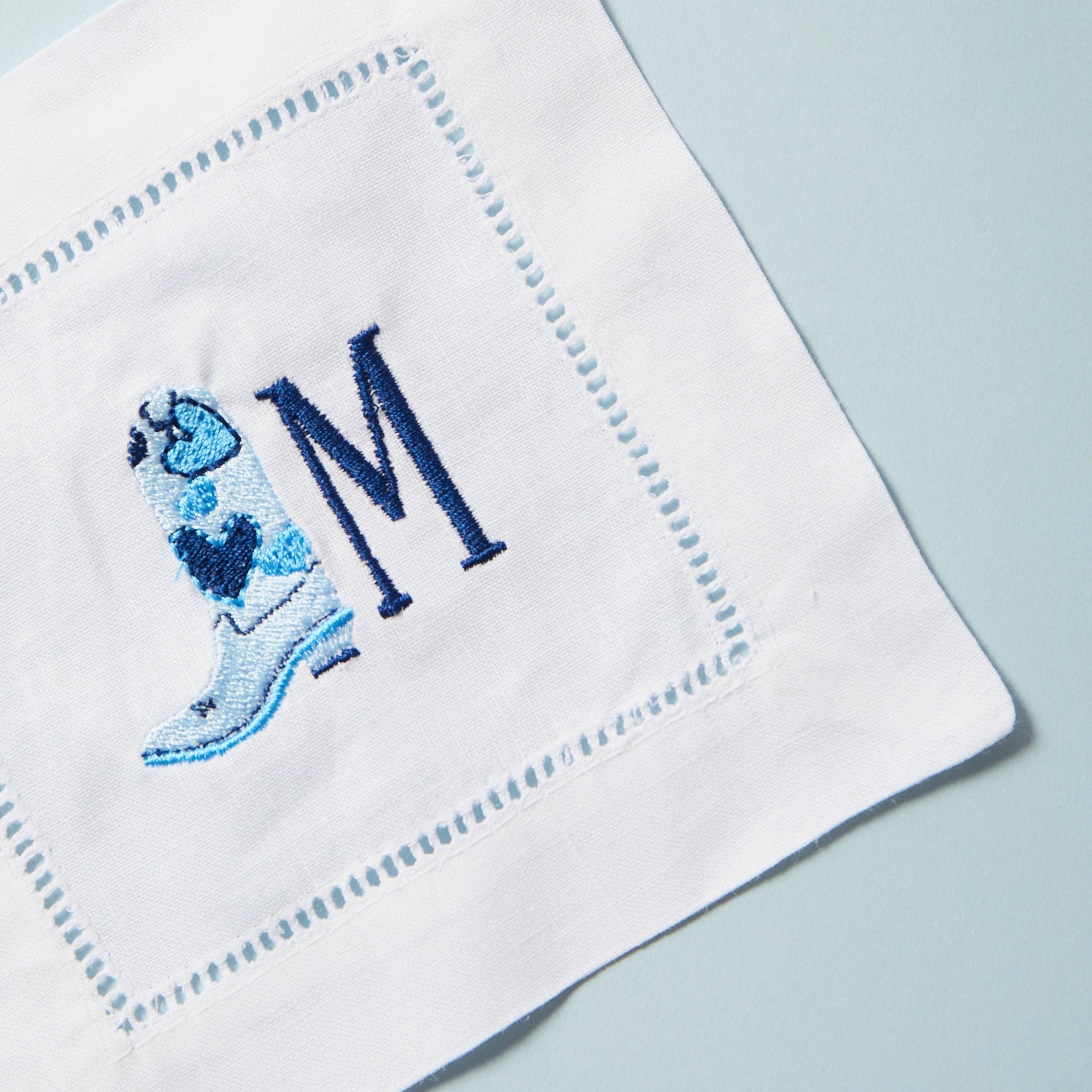 A cocktail napkin with an embroidered blue cowboy boot and navy initial.