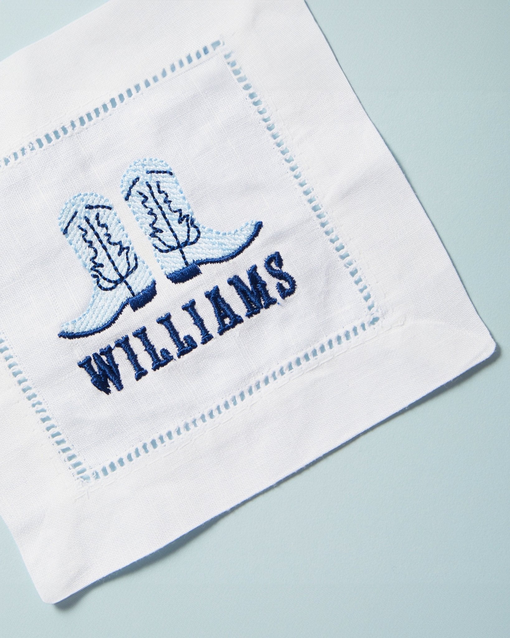 An embroidered cocktail napkin with a pair of blue cowboy boots and a custom name.