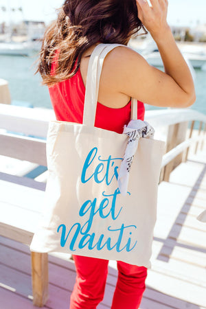 A woman in a red jumpsuit shows off a cotton tote bag which reads "let's get nauti" in a blue font on a dock.