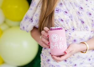 A woman holds up a pink can cooler with "Oh Baby" printed on it in white.