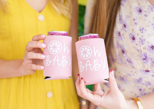 Two women hold up pink can coolers with "Oh Baby" printed on them in white.