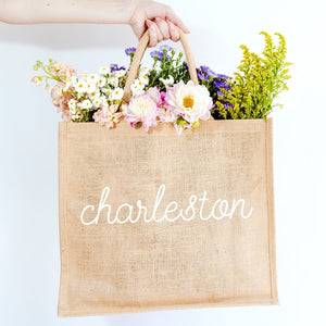 A person holds a jute tote which is customized with "Charleston" in a white cursive font.
