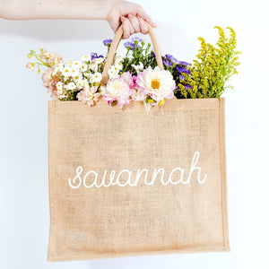 A person holds a jute tote which is customized with "savannah" in a white cursive font.