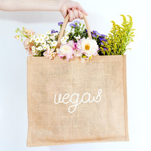 A person holds a jute tote which is customized with "vegas" in a white cursive font.