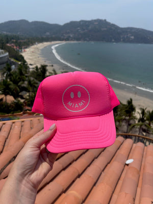A woman holds a hot pink trucker hat with a smiley face that says "Miami"