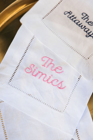 A cocktail napkin that is embroidered in light pink font with a custom last name.