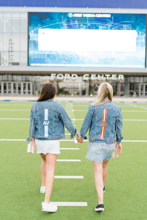 Two women walking on a field sport their striped monogram jean jackets.