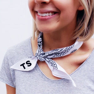 A person wears a white bandana around her neck with a black monogram.