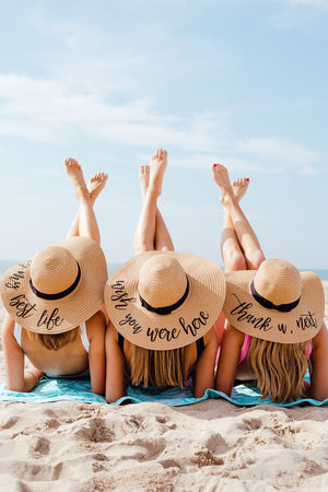 A group of women on the beach wear customized floppy hats with fun phrases printed on them.