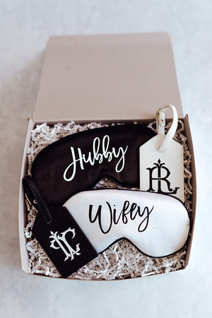 A box is opened to reveal a black and white "Hubby" and "Wifey" sleep mask with matching black and white monogrammed luggage tags