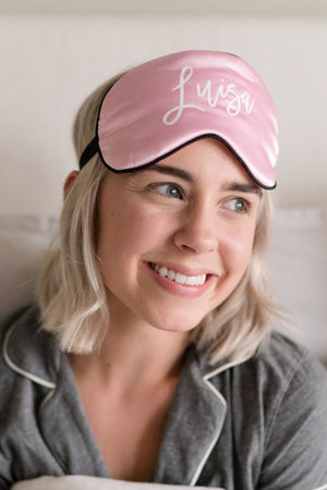 A woman wears a light pink sleep mask personalized with her name on it.