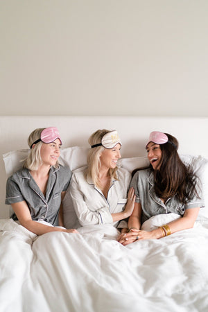 Three women lay in bed laughing with each other and wearing custom sleep masks.