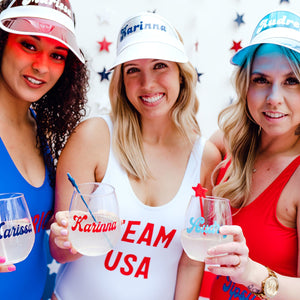 Three women hold up their wine glasses customized with their names while decked out in red, white, and blue celebration attire.