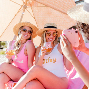 Two girls sit drinking out of their stemless wine glasses with a retro monogram and wearing custom swimsuits.
