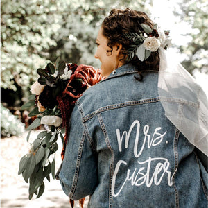 A bride holds some flowers and wears a customized jean jacket with her new last name and a veil.