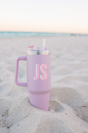 A pink, monogrammed tumbler sits in the sand on a beach