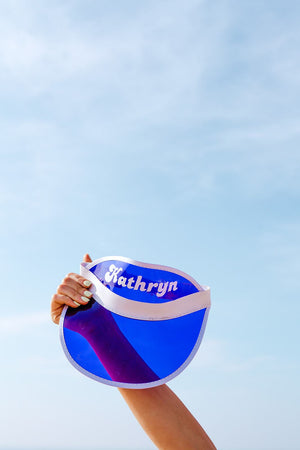 A person holds up their purple visor with a custom name on it up to the sky