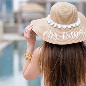 A woman at a pool grabs onto her custom floppy beach hat which reads "Mrs. Dillon" in white.