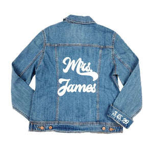 A jean jacket is laid down showing the customized name across the back and custom date attached to the cuff.